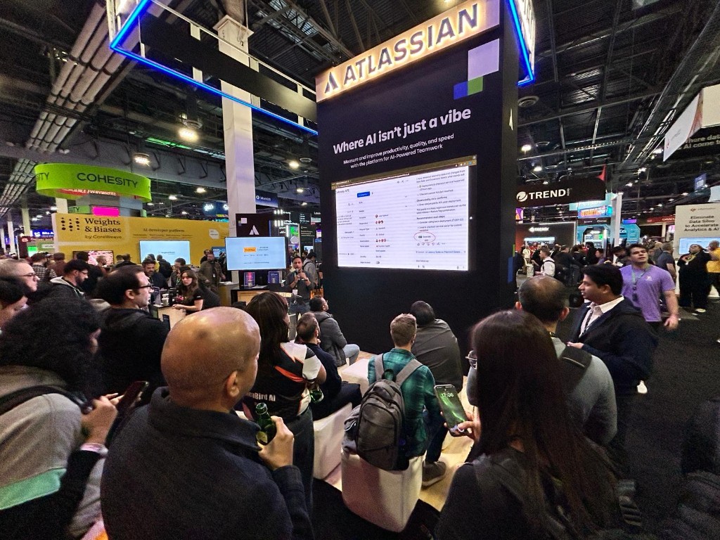 Wide shot of the Atlassian booth at AWS re:Invent 2025 with a crowd gathered, set against the AWS show floor with Cohesity, Weights & Biases, and Trend Micro booths visible nearby.