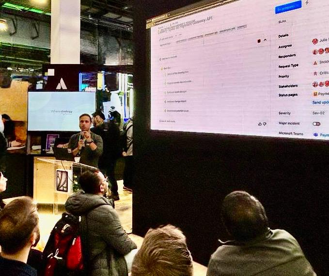 Dushyant Sharma speaking with a handheld mic at AWS re:Invent 2025, with a large screen showing a Jira Service Management incident view and a smaller monitor displaying a strategy slide.
